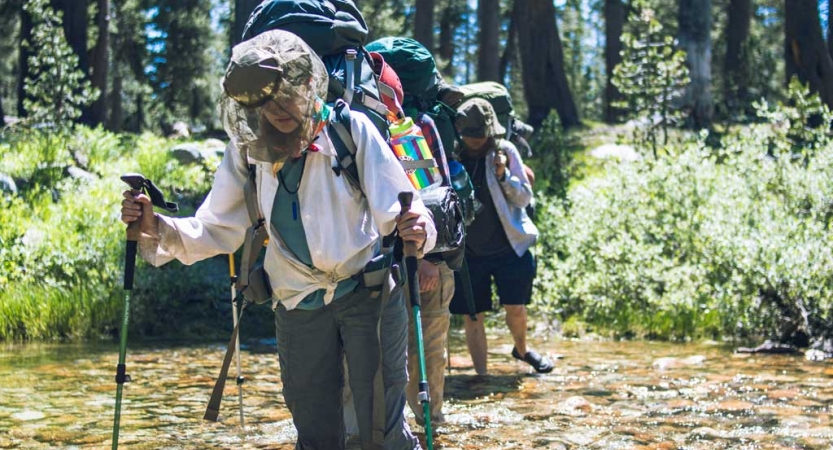A group of people wearing backpacks and mosquito nets use trekking poles to cross a shallow creek. 